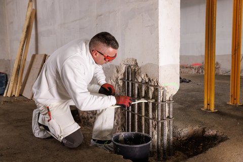 Arbeiter trägt graue Masse auf freigelegte Stahlbewehrung am Pfeiler auf.